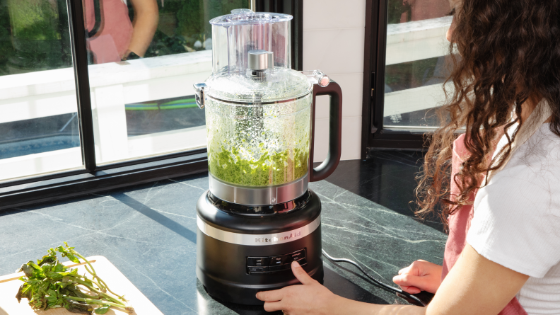 A person presses the button on the KitchenAid Food Processor to process a mixture of green herbs.