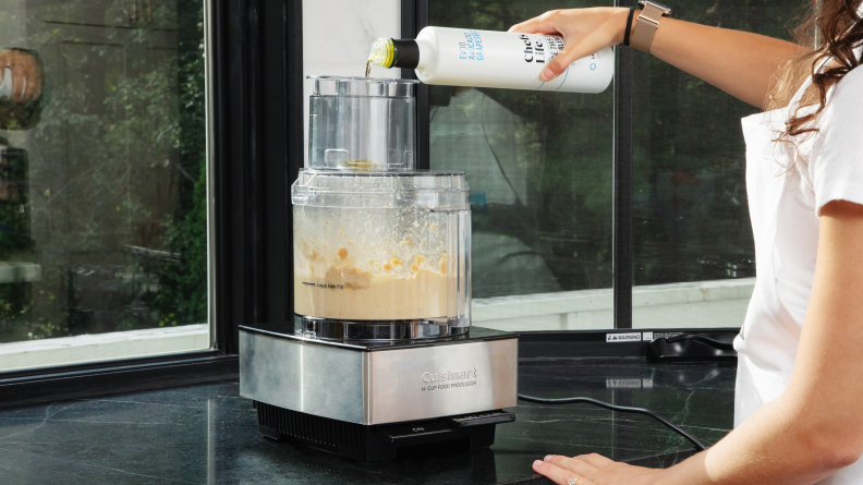 A person pouring olive oil into a Cuisinart food processor while blending hummus.